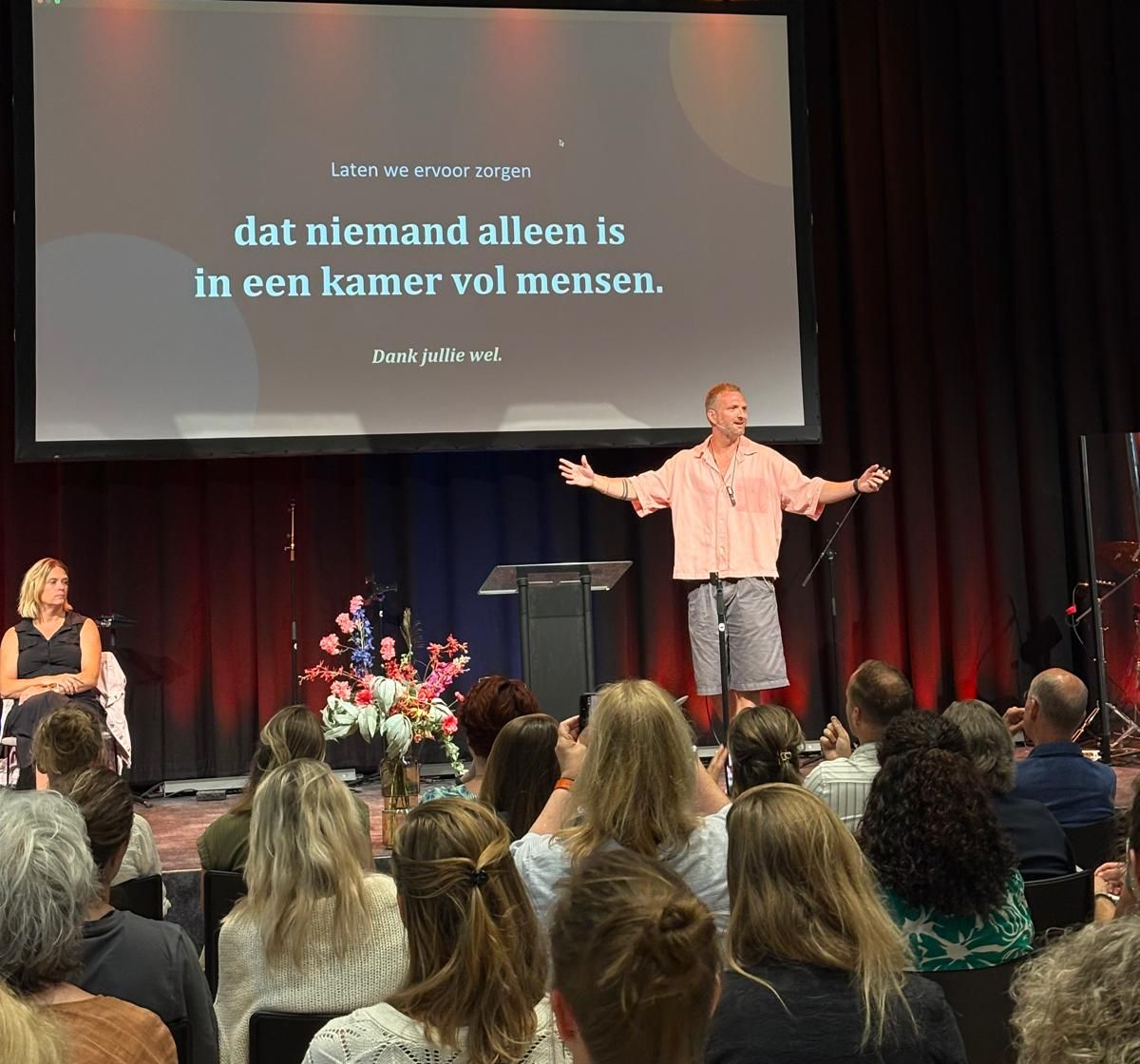 Pieter staat met gespreide armen op een podium voor een volle zaal; op het scherm achter hem staat: dat niemand alleen is in een kamer vol mensen